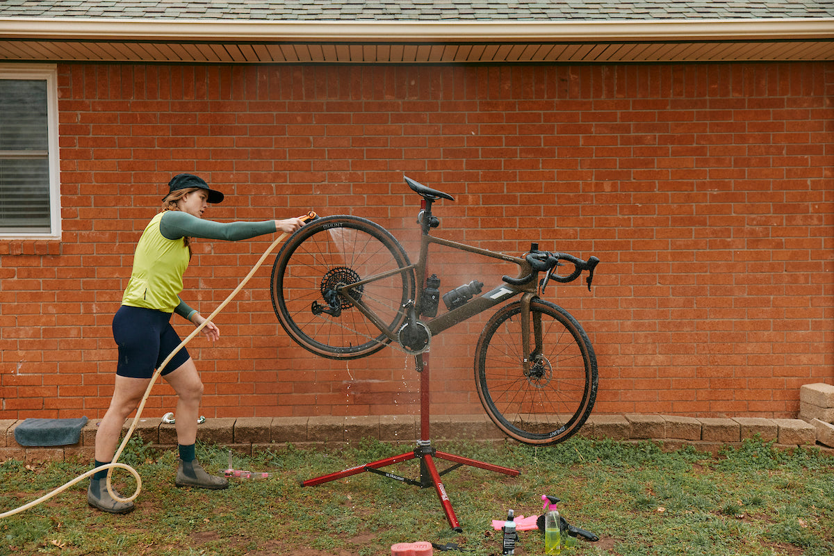 Woman using hose to spray bike in repair stand outdoors next to building.