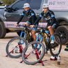 Two young mountain bike racers on mountain bikes warming up with bikes mounted on Omnium stationary trainers outdoors.