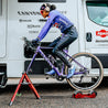 Cyclocross racer on bike warming up on Omnium stationary trainer next to team truck under tent.